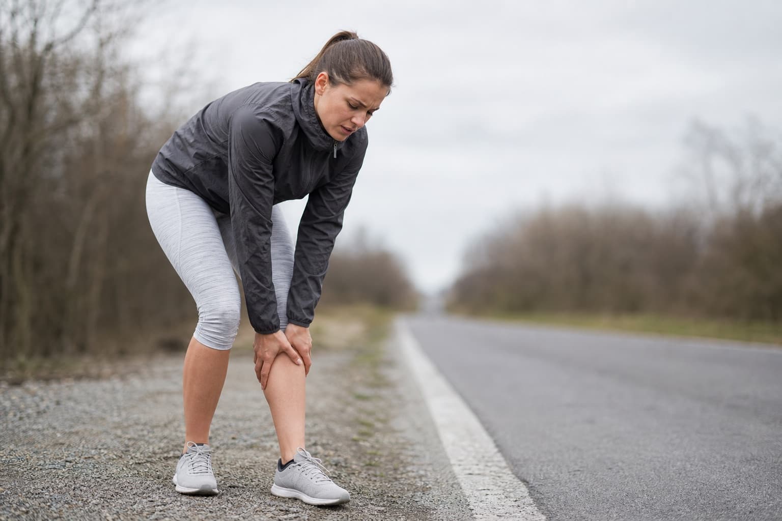 Runner paused on a road, hand on a sore lower leg with a cautious expression.