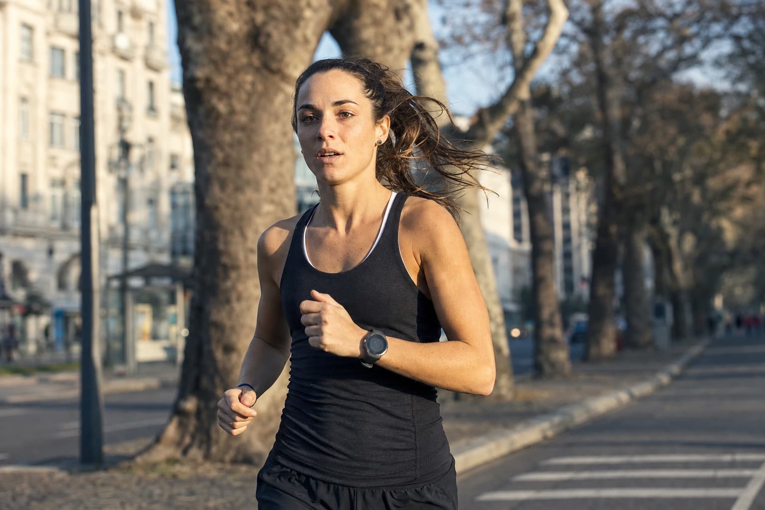 Runner mid-stride wearing a GPS watch on a tree-lined city street.